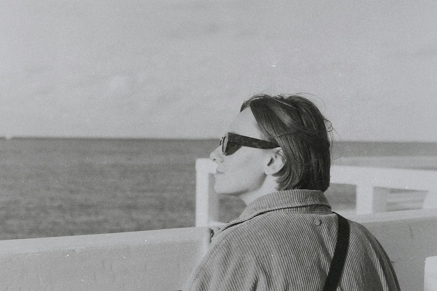 Person standing at a waterfront railing, looking out toward the open sea on a calm, overcast day