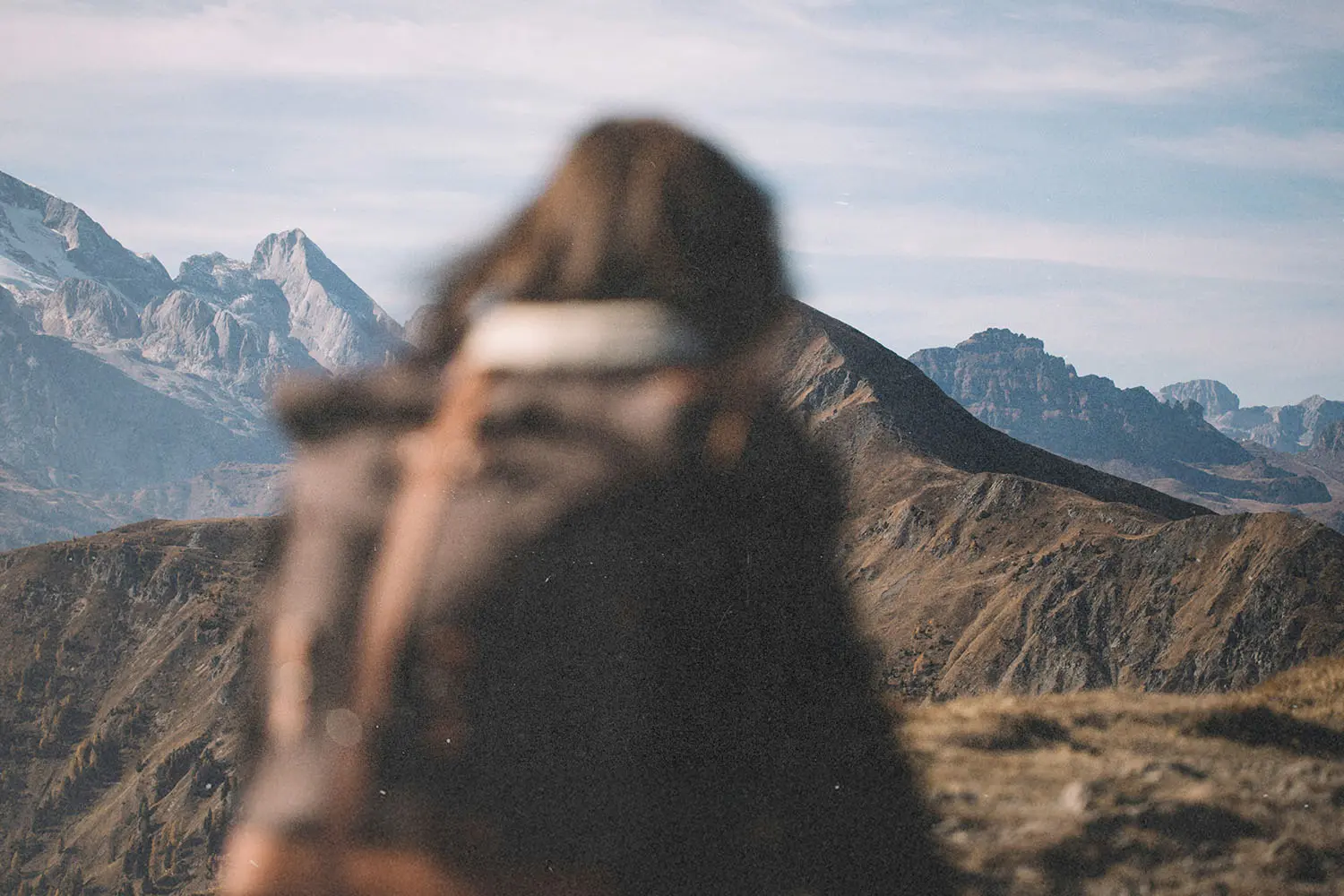 A person with a backpack stands on a mountain trail, facing a distant range of rugged peaks