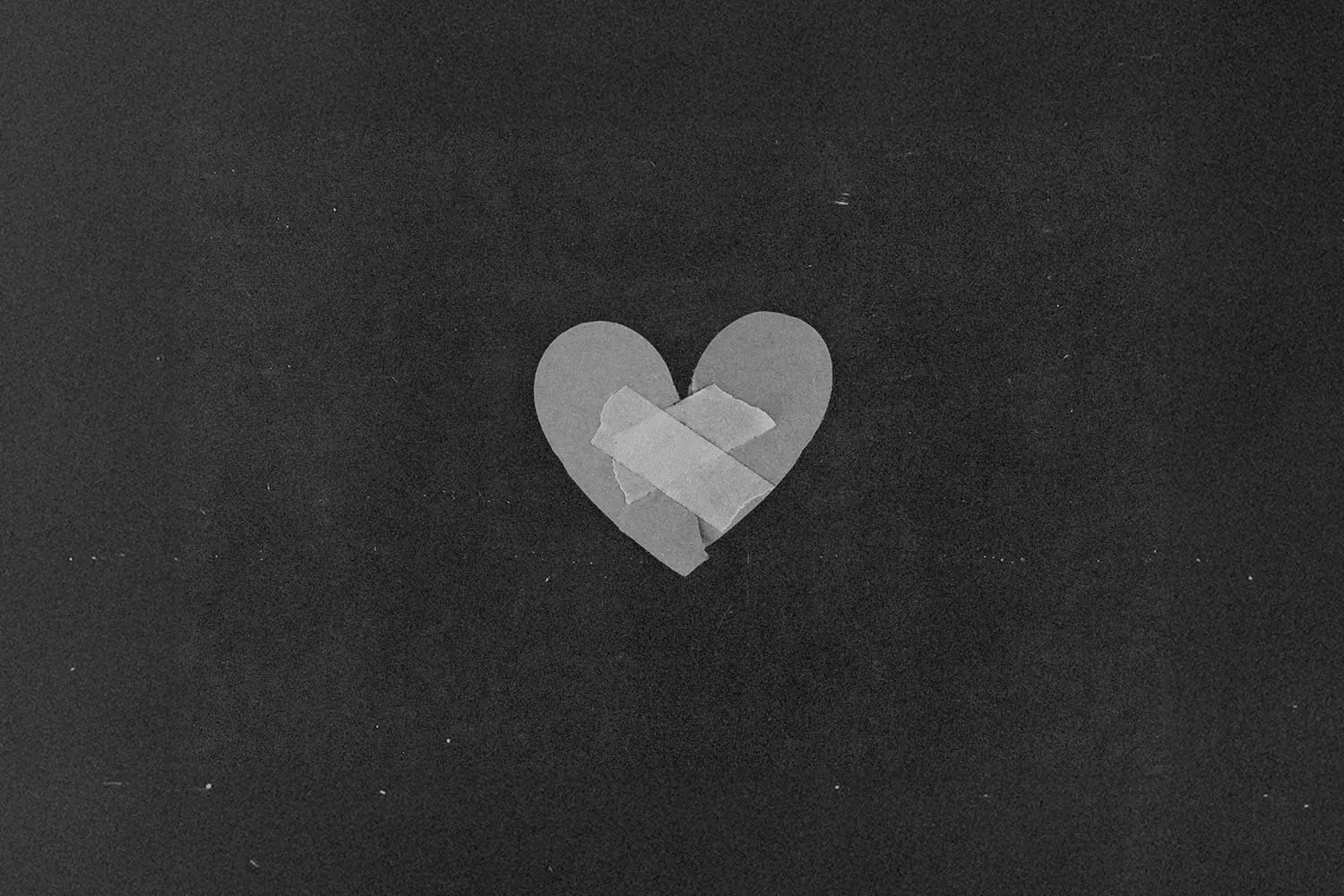 A simple paper heart sits against a dark background with two pieces of tape placed in an “X” shape across the center