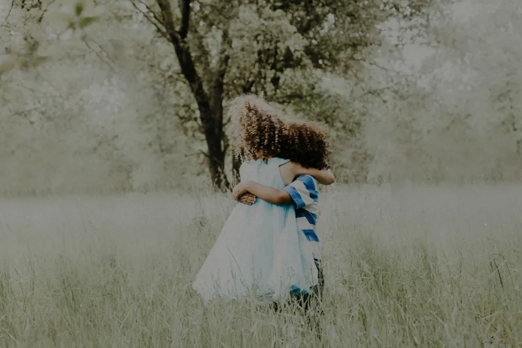 Two children embrace while standing in tall grass beneath trees in a softly lit outdoor setting