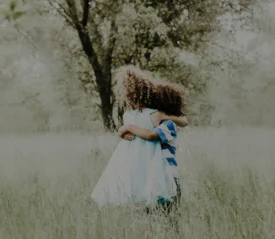 Two children embrace while standing in tall grass beneath trees in a softly lit outdoor setting