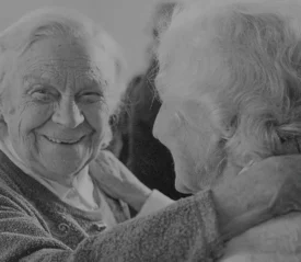 Two older adults embrace closely, one placing a hand gently on the other’s shoulder