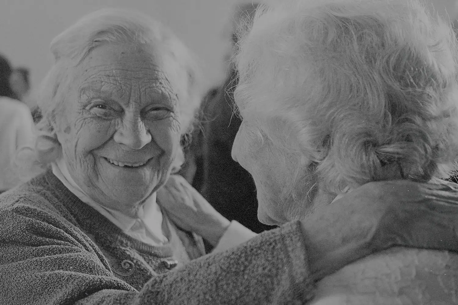Two older adults embrace closely, one placing a hand gently on the other’s shoulder
