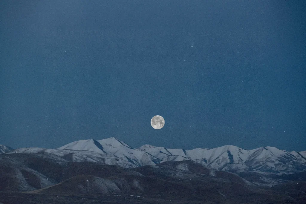 Full moon rising above a snow‑covered mountain range under a deep blue sky