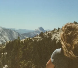 Woman with windblown hair looking out over a mountain landscape filled with pine trees and rugged cliffs