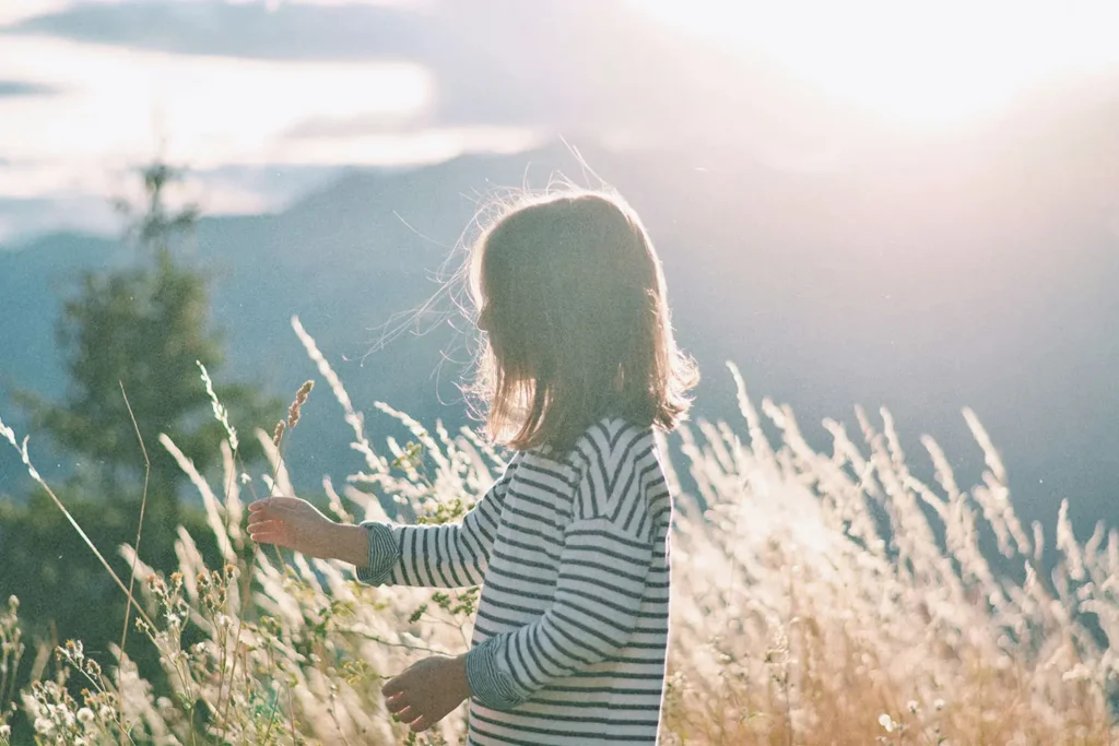 A child stands in a sunlit field of tall grass, gently touching the plants as the mountains glow softly in the background
