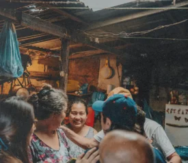 Several people gather around someone in a rustic, dimly lit shelter, placing comforting hands on their shoulders