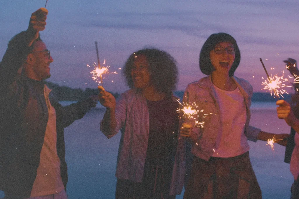 A group of people stand together at dusk near the water, holding sparklers that glow against the purple sky