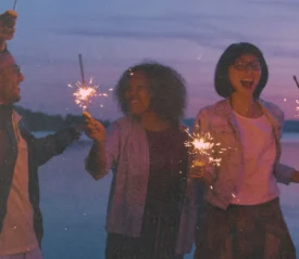 A group of people stand together at dusk near the water, holding sparklers that glow against the purple sky