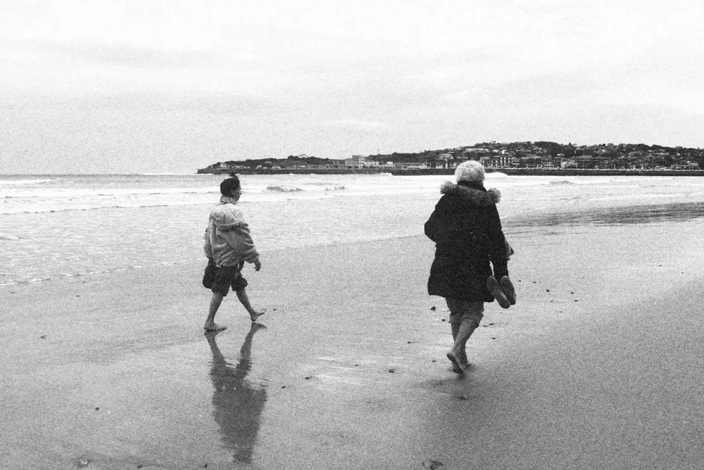 Two people walking barefoot along a quiet beach shoreline on a cloudy day, with a town visible in the distance