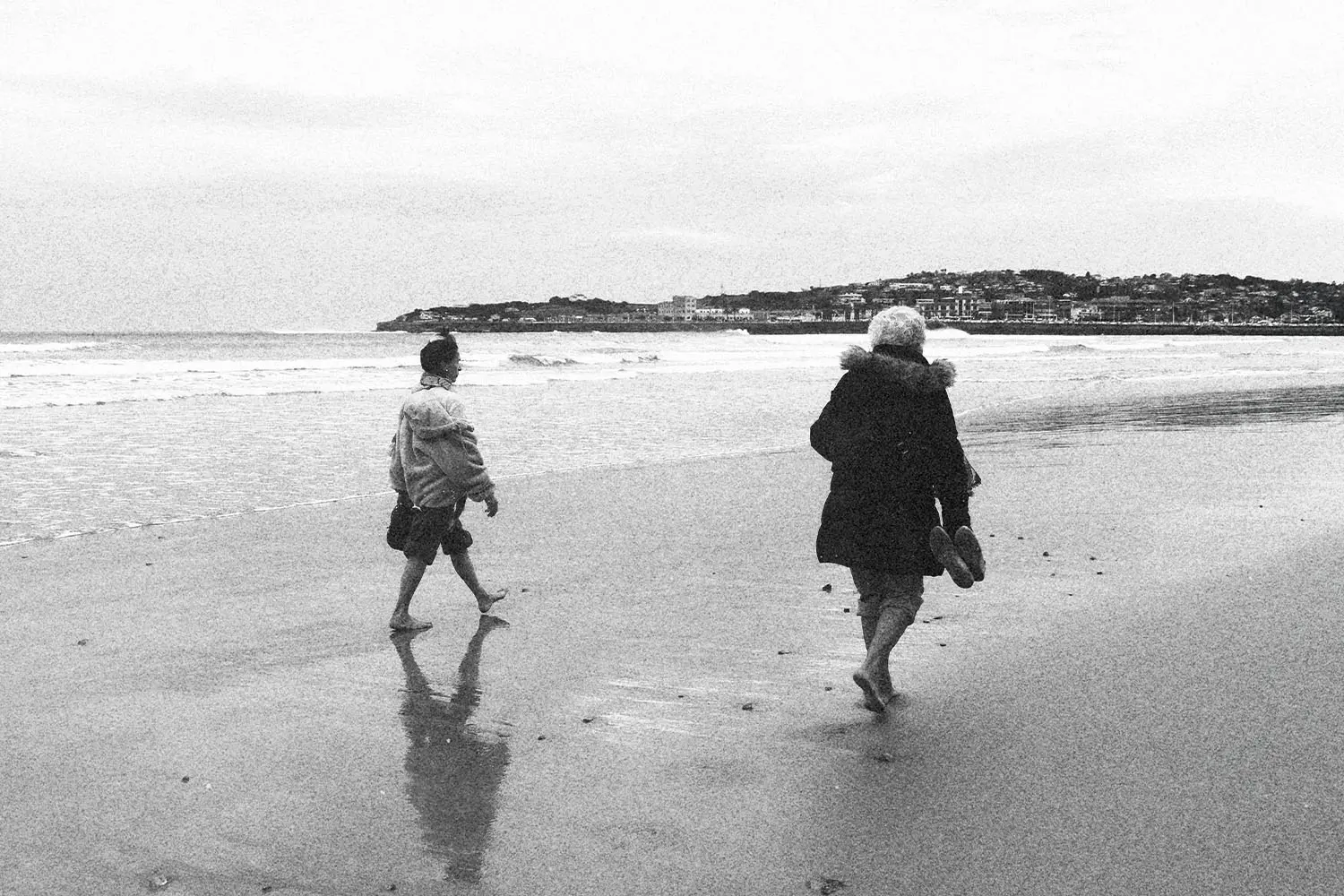 Two people walking barefoot along a quiet beach shoreline on a cloudy day, with a town visible in the distance