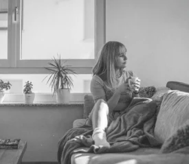 Person relaxing on a sofa with a blanket and holding a warm drink near a bright window with potted plants