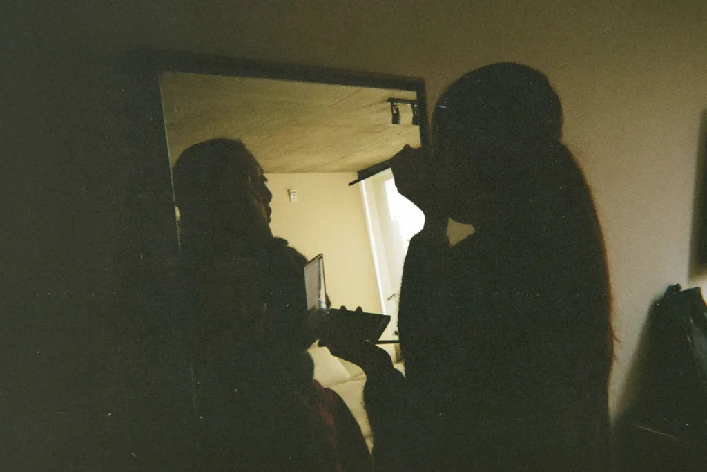 Person applying makeup in front of a mirror in a dimly lit room, holding a compact and brush