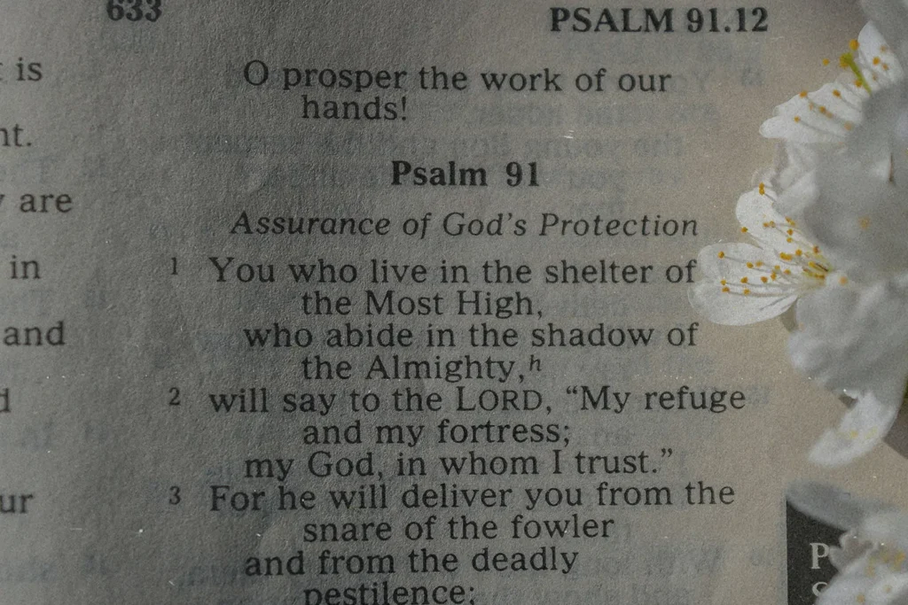 A close-up view of an open Bible showing the beginning of Psalm 91, with small white flowers resting along the page