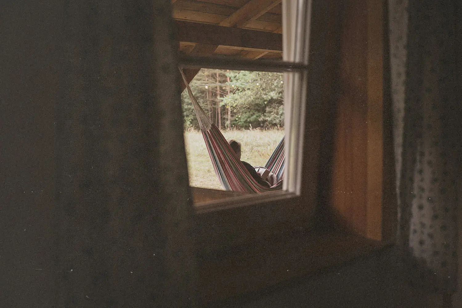 View through a window of a person reclining in a striped hammock under a wooden porch, with trees and grass in the background