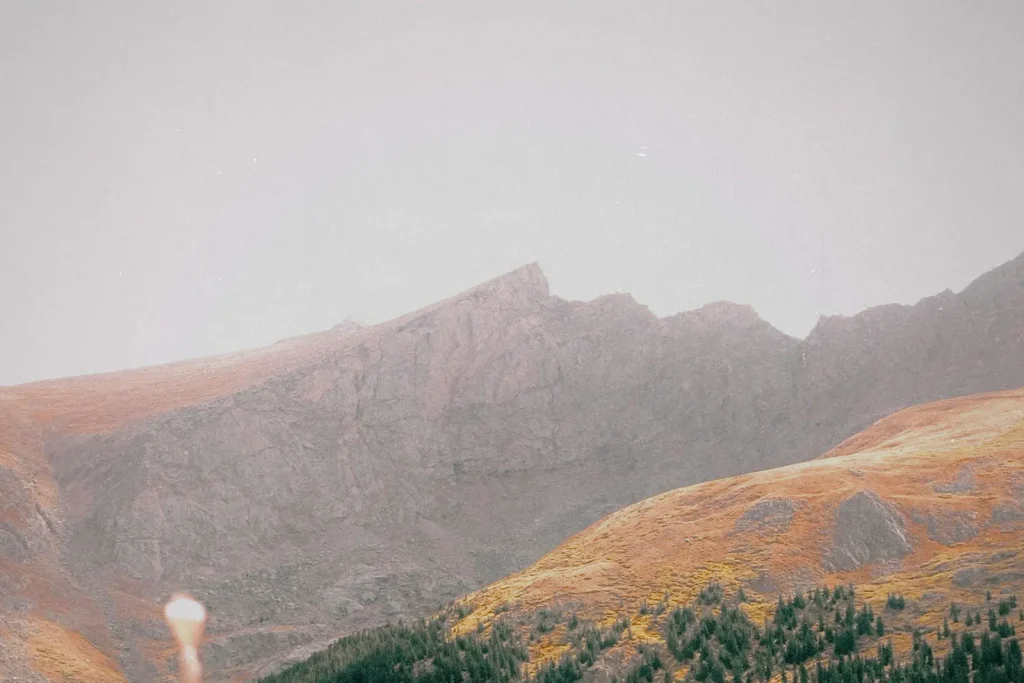Rugged mountain landscape with steep rocky slopes rises beneath a hazy sky, with autumn-colored grass and scattered trees in the foreground