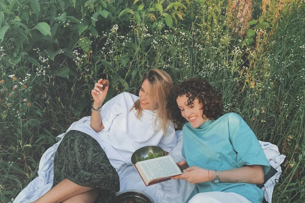 Two people relax on a blanket in a grassy, flower‑filled meadow, sharing fruit and reading a book together