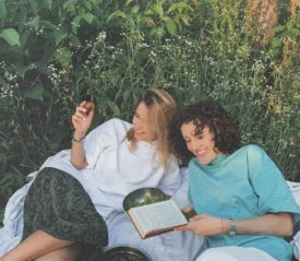 Two people relax on a blanket in a grassy, flower‑filled meadow, sharing fruit and reading a book together