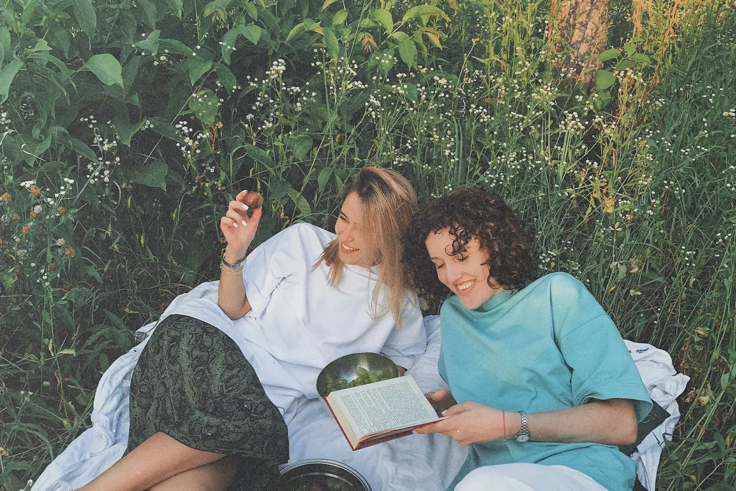 Two people relax on a blanket in a grassy, flower‑filled meadow, sharing fruit and reading a book together