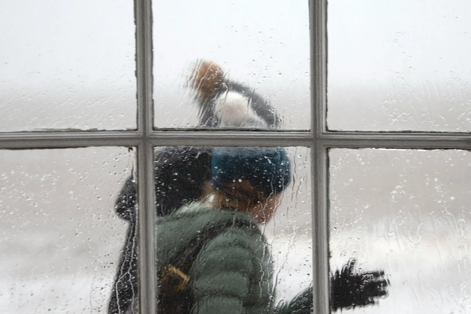 Two people in winter clothing walk past a rain‑covered window, their figures softened by the water droplets on the glass