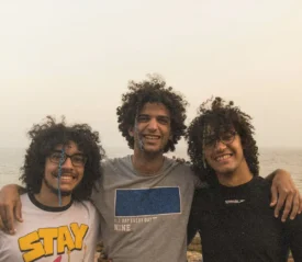 Three friends stand arm in arm near the shoreline, with the sea and sky forming a soft backdrop