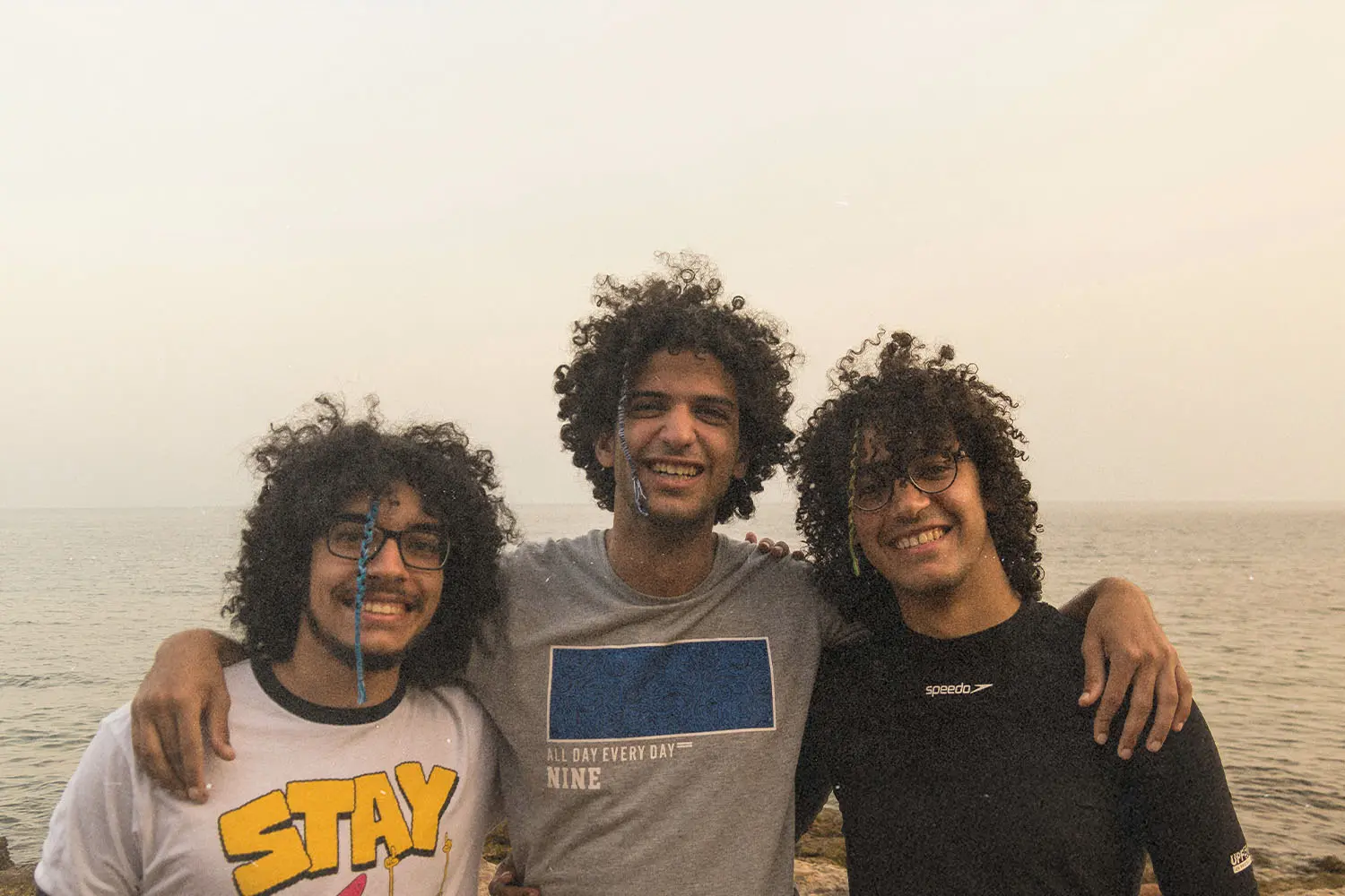 Three friends stand arm in arm near the shoreline, with the sea and sky forming a soft backdrop
