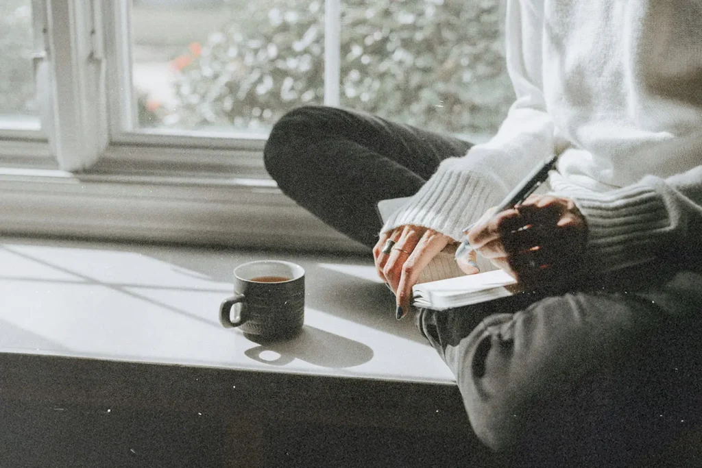 Person sitting on a sunny windowsill with a notebook and a cup of coffee, writing in soft natural light