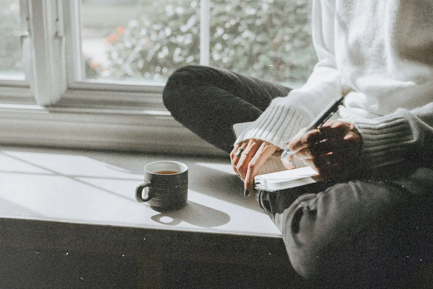 Person sitting on a sunny windowsill with a notebook and a cup of coffee, writing in soft natural light