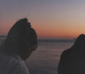 Two people stand near the water at sunset, their silhouettes softly outlined against a warm orange and purple sky