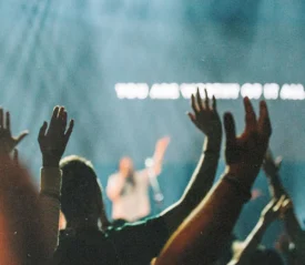 Worship crowd raising their hands during a live praise event with stage lights and lyrics glowing on a screen in the background