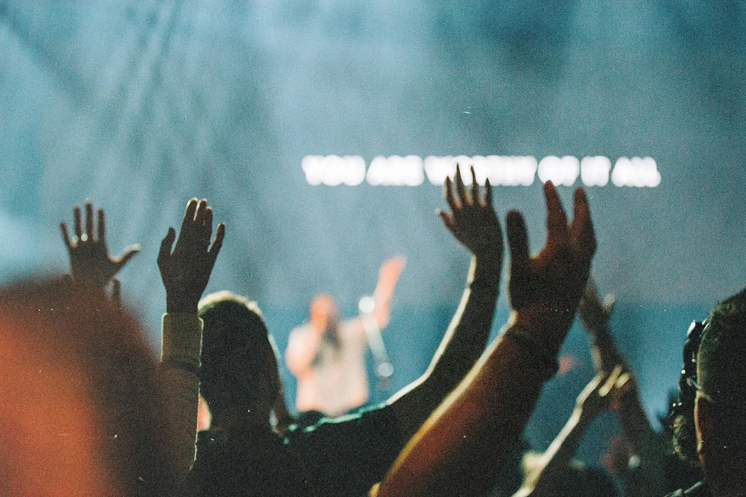 Worship crowd raising their hands during a live praise event with stage lights and lyrics glowing on a screen in the background