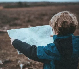 A child wearing a blue jacket stands outdoors holding a large map, looking out over a wide, open landscape