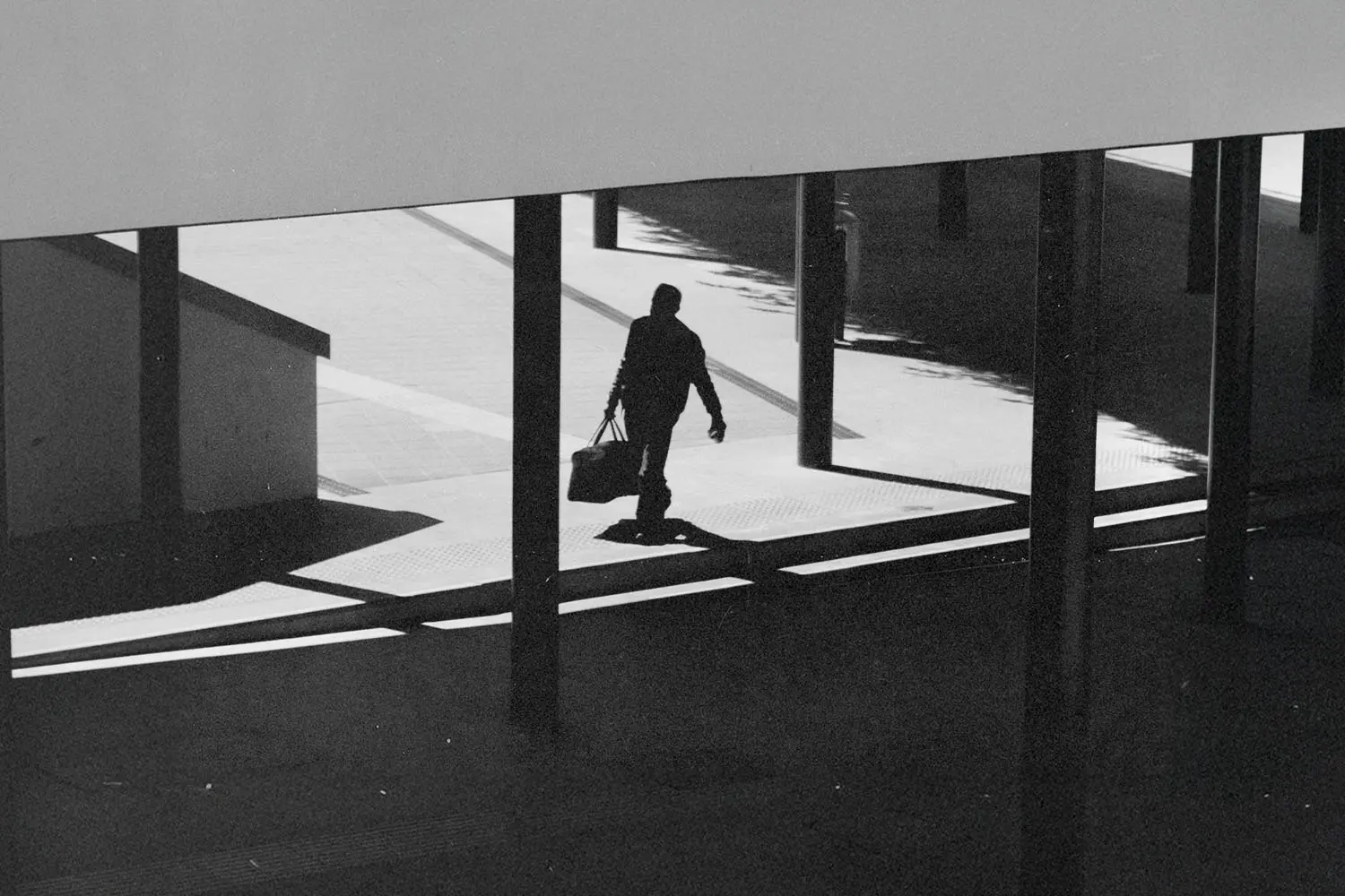 A silhouetted figure walks through a modern outdoor walkway carrying a bag, with strong shadows cast across the ground