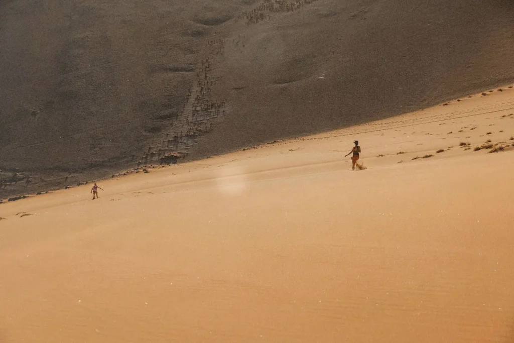 Two people walk across a vast desert slope, appearing small against the large sandy landscape