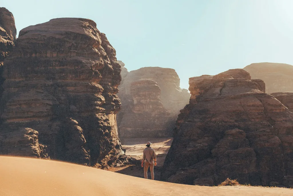Person standing on a sandy desert path surrounded by towering rock formations under a bright sky
