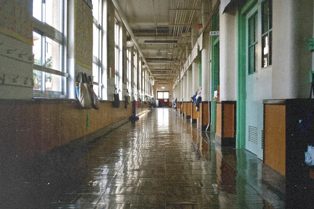 A long, empty school hallway lined with windows and classroom doors reflects bright light on its shiny floor