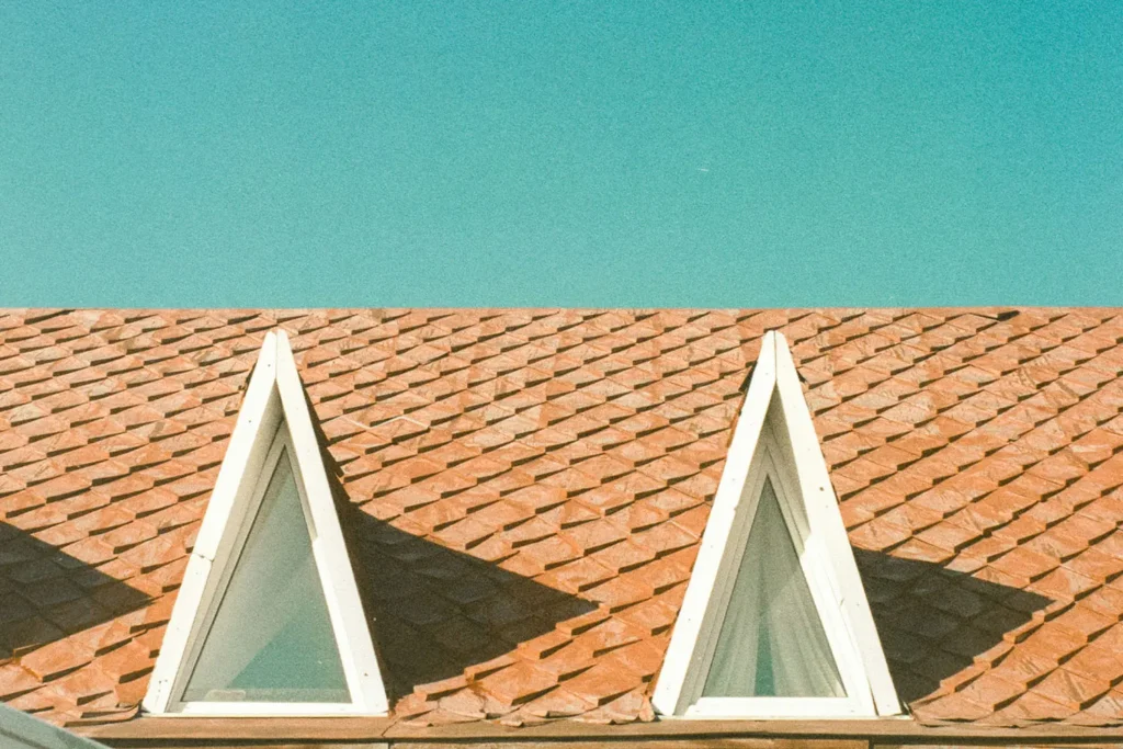 Two triangular attic windows on a red shingle roof beneath a clear blue sky