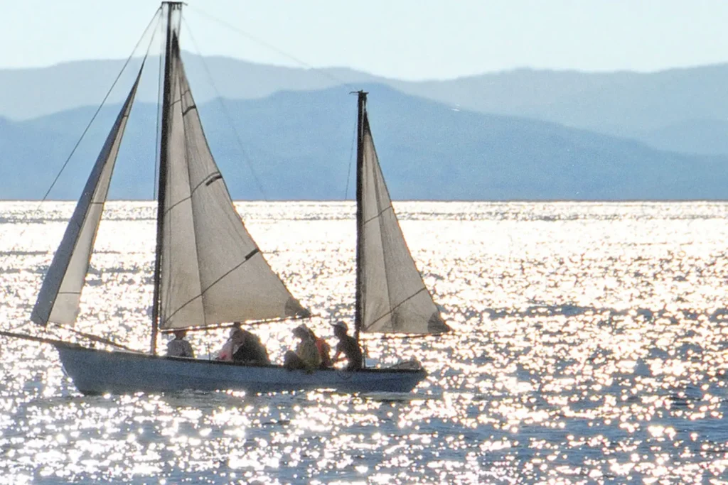 A small sailboat glides across shimmering sunlit water, carrying several people beneath three raised sails, with distant mountains visible on the horizon