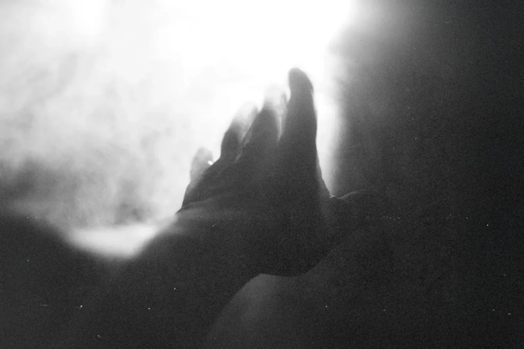 Hand reaching toward a bright light surrounded by soft, glowing mist in a black‑and‑white scene