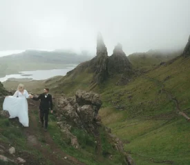 ride and groom holding hands while hiking through mist-covered highlands with steep cliffs and rolling green hills