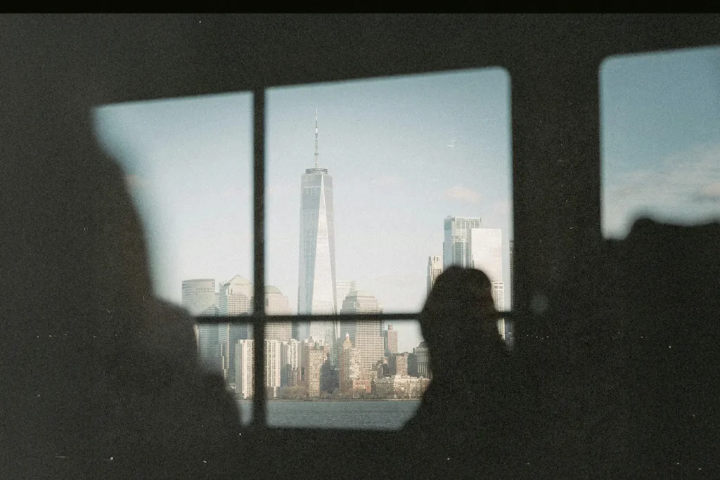 City skyline framed through a window, featuring a tall modern skyscraper and surrounding buildings with silhouetted observers in the foreground