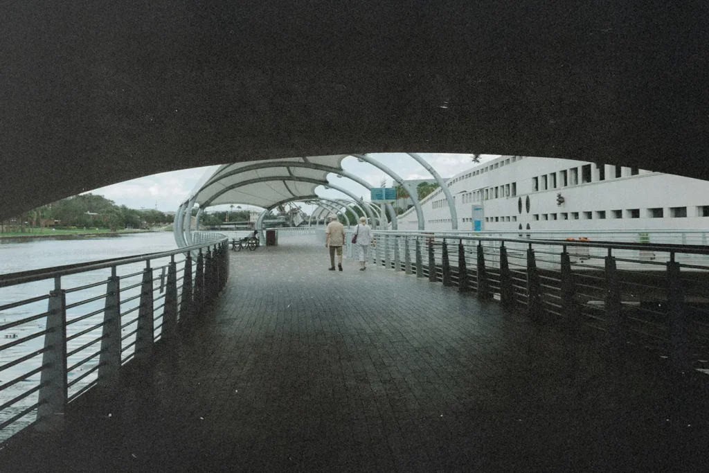Two people walk together along a covered riverside boardwalk with railings on both sides