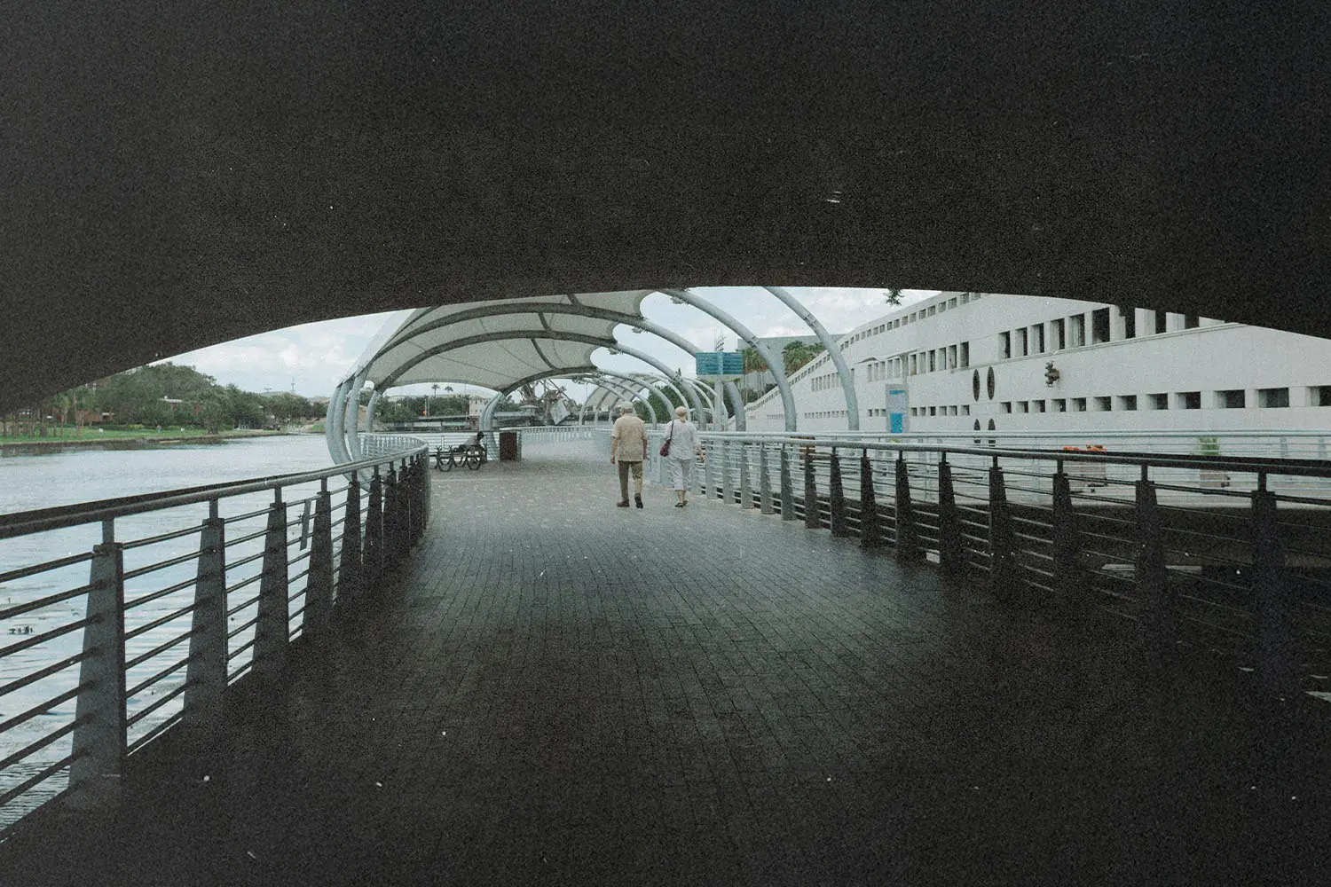 Two people walk together along a covered riverside boardwalk with railings on both sides