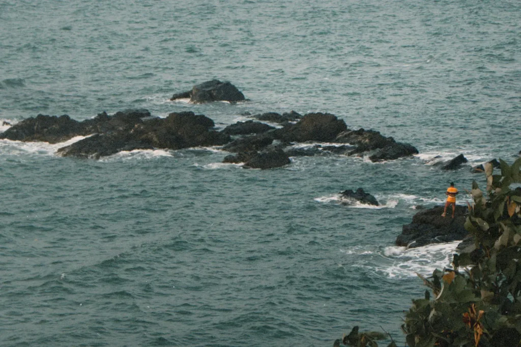 A person wearing a yellow shirt stands on dark rocks surrounded by ocean waves