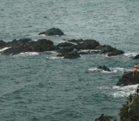 A person wearing a yellow shirt stands on dark rocks surrounded by ocean waves
