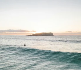 Two surfers sitting on their boards in gentle ocean waves at sunrise with a small island in the distance
