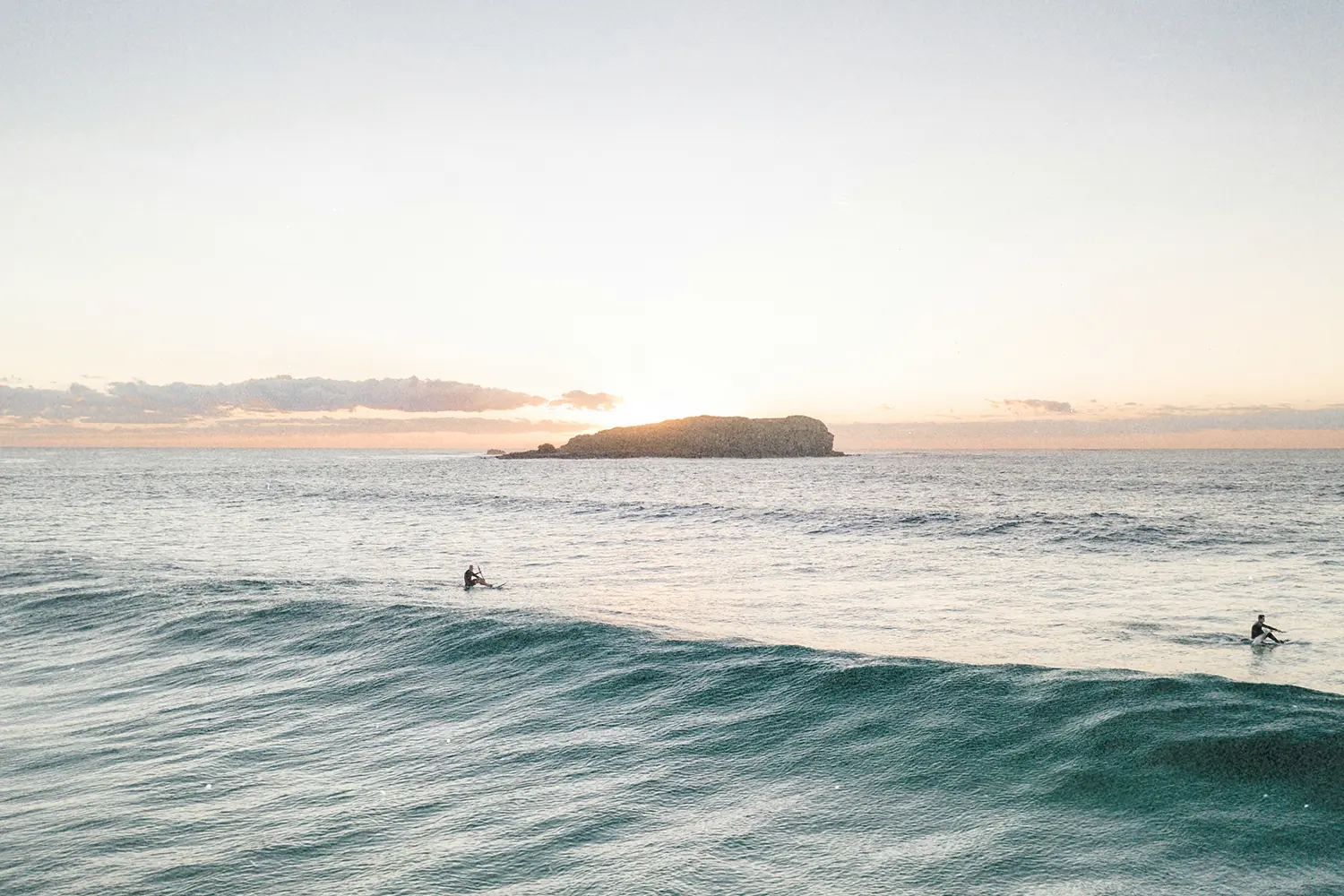 Two surfers sitting on their boards in gentle ocean waves at sunrise with a small island in the distance