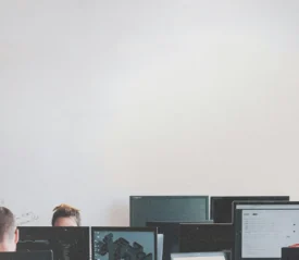 Several people sit at desks working on computers in a bright room with a large blank wall above them