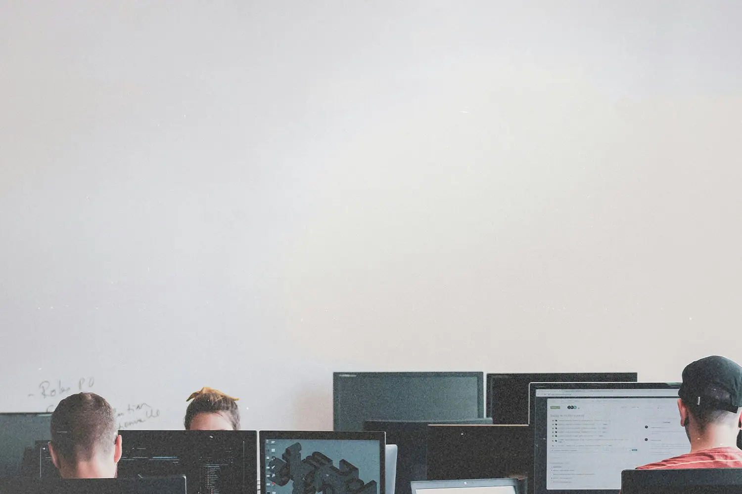 Several people sit at desks working on computers in a bright room with a large blank wall above them