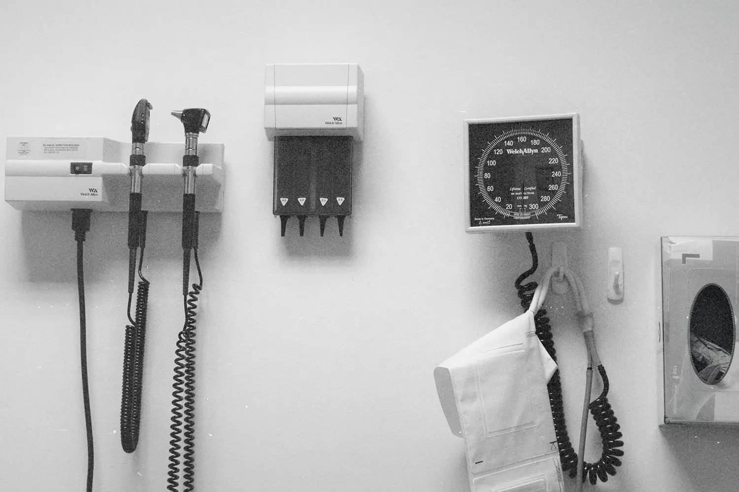 Wall-mounted medical equipment, including an otoscope, blood pressure gauge, and storage units, arranged neatly in a clinical exam room